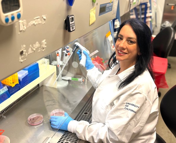 A scientist wearing a white lab coat and blue gloves works in a lab. She smiles while using a pipette near petri dishes.