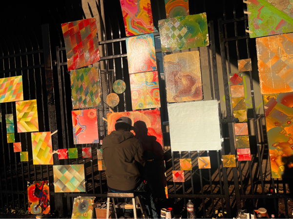 Painter working on a piece on a city fence, other art hung up around him