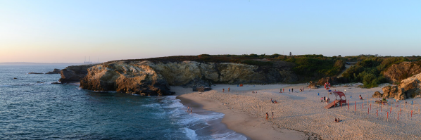A wide view of a sandy beach bordered by rocky cliffs, with small groups of people walking and sitting near the shoreline as waves roll in at sunset.