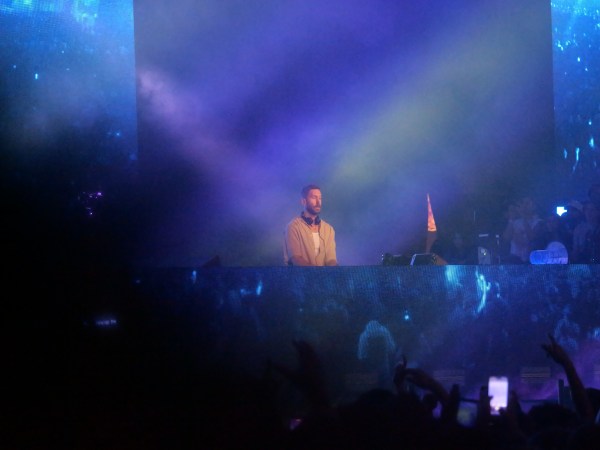 Calvin Harris performs during a live set amid blue and purple stage lighting, as fans raise their hands in the crowd below. (Courtesy of Sebastien dePierre)
