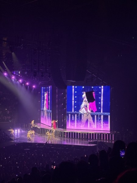 Performer in a bright pink outfit on a large concert stage with dancers, purple lighting, and giant video screens showing the performance.
