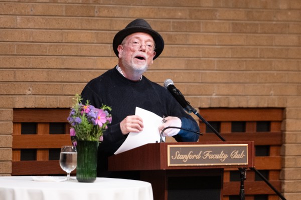 D.A. Powell glances up from a packet of poems to speak to the audience from behind the podium at Stanford Faculty Club, near a table with flowers and a glass of water.