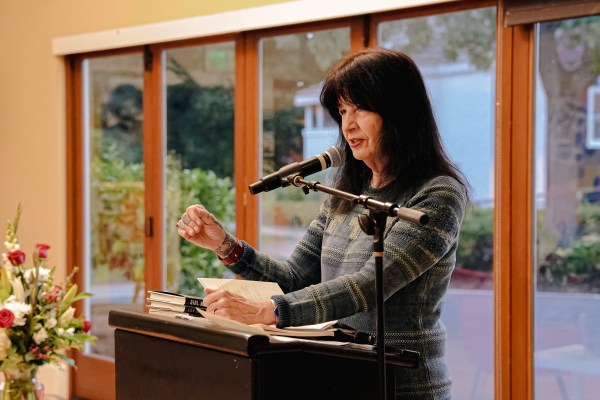 Joy Harjo reads into a microphone mounted on a podium in front of a panel of windows.