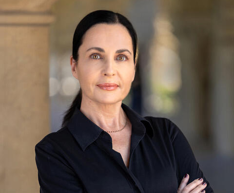 Woman with dark hair in ponytail, wearing a black blouse, stands with arms crossed.