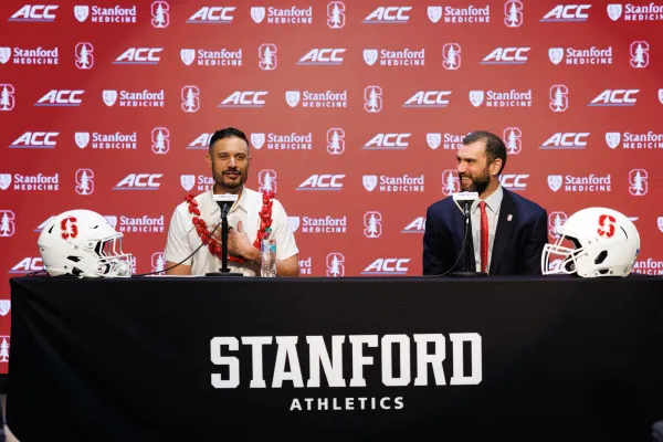 Two men sitting at press conference table with red screen behind them.