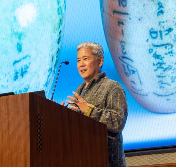 Hideo Mabuchi speaking on a lectern, with a photo of ceramics projected in the background