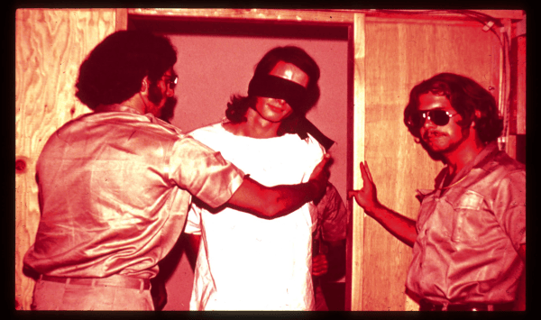A photo of a blindfolded "prisoner" with "guards" during the Stanford Prison Experiment.