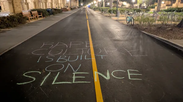 Picture of a lamp-lit Lasuen Mall road besides Stanford Graduate School of Education, with chalking on ground that reads: “RAPE CULTURE IS BUILT ON SILENCE”